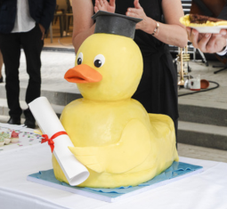 Large, 3D cake shaped like a duck wearing a graduation cap and holding a rolled up degree.
